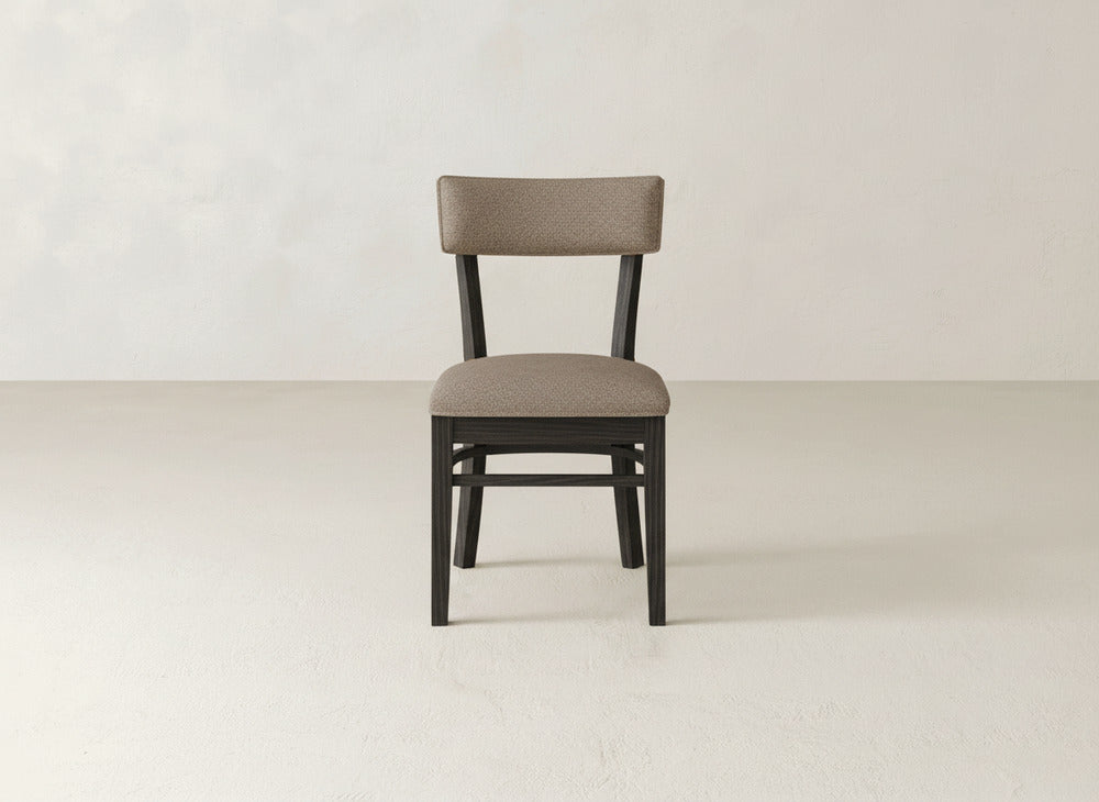The Everly Side Dining Chair by James+James, featuring a padded beige fabric seat and backrest, is centered against a plain, light-colored wall and floor.
