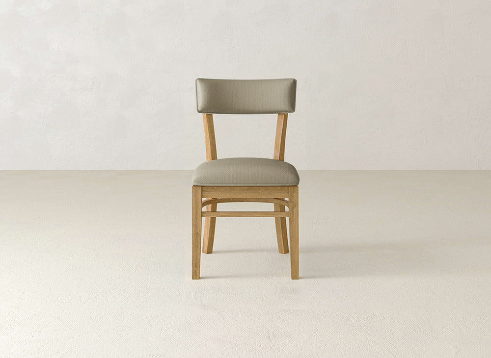 The Everly Side Dining Chair by James+James, with light brown wooden legs and frame, features a padded light gray seat and matching backrest. It’s shown against an off-white wall on a light-colored floor.