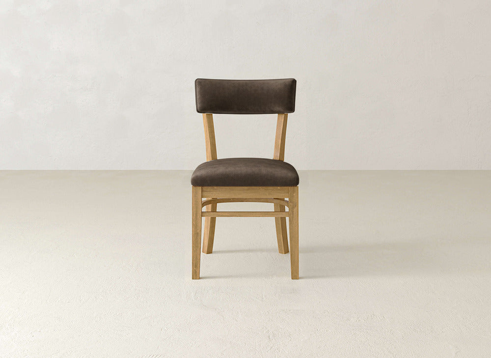 The Everly Side Dining Chair by James+James, featuring a dark brown cushioned seat and backrest, is centered against a plain, light-colored wall and floor.