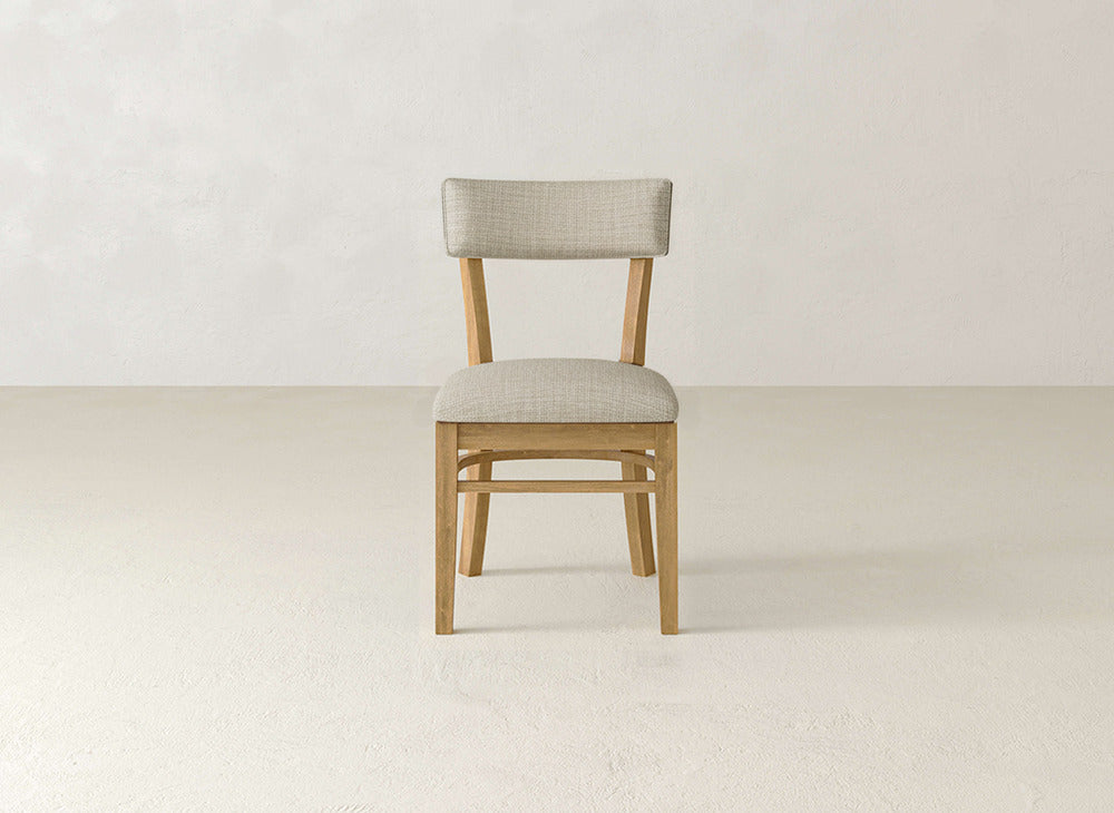 The Everly Side Dining Chair by James+James features a light beige upholstered seat and backrest, set on a plain, light-colored floor against a smooth off-white wall.