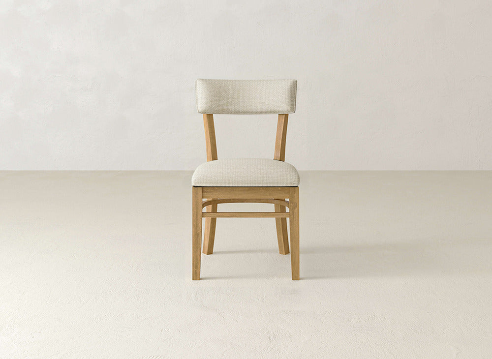 The Everly Side Dining Chair by James+James features a light cushioned seat and back, centered on a textured light floor against an off-white wall, with its mid-century design facing forward.
