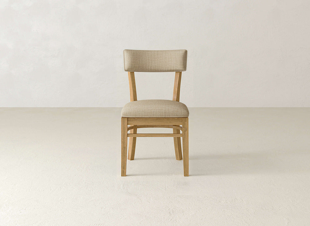 The Everly Side Dining Chair by James+James, featuring a light beige cushioned seat and backrest, is centered on a light floor against an off-white wall and viewed from the front.