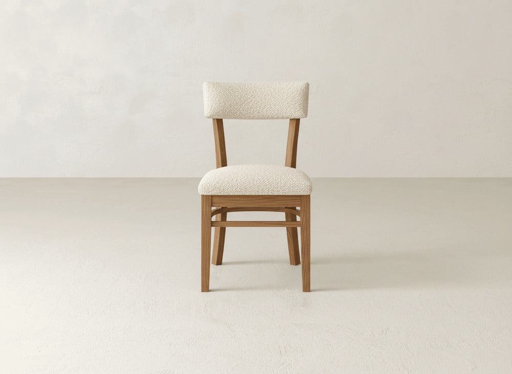 The Everly Side Dining Chair by James+James, featuring a light beige cushioned seat and backrest, sits centered in a minimalist room with pale floors and walls.