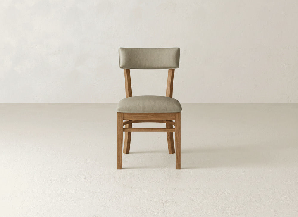 The Everly Side Dining Chair by James+James, featuring a cushioned seat and backrest in light beige fabric, is centered against a plain, light-colored wall and floor.