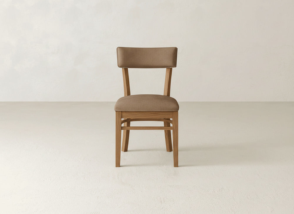 The Everly Side Dining Chair by James+James, featuring a light brown cushioned seat and backrest, is centered and facing forward against a plain, light-colored wall and floor.