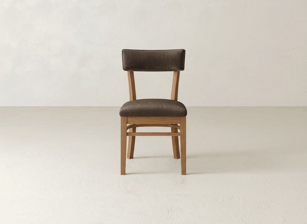 The Everly Side Dining Chair by James+James, featuring a dark brown cushioned seat and backrest, is showcased in the center of a minimal, light-toned room with a plain background.