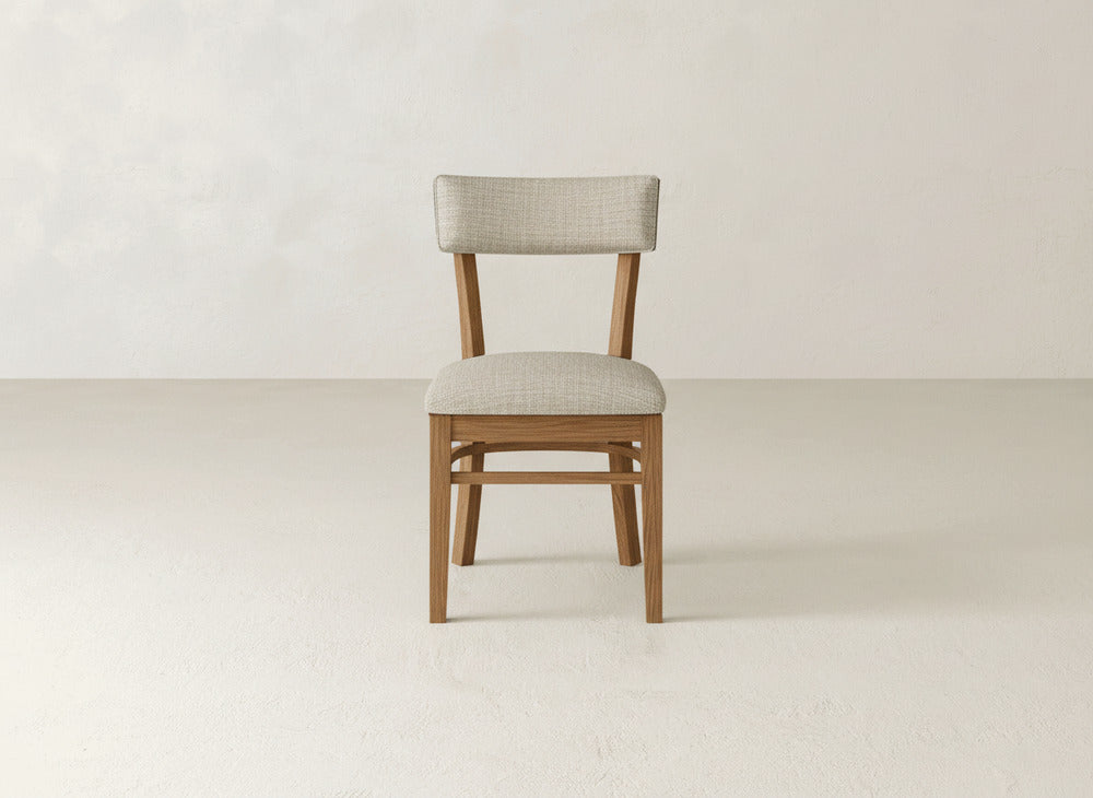 The Everly Side Dining Chair by James+James showcases a wooden frame with light beige upholstery on the seat and backrest, set against a plain off-white wall and floor.