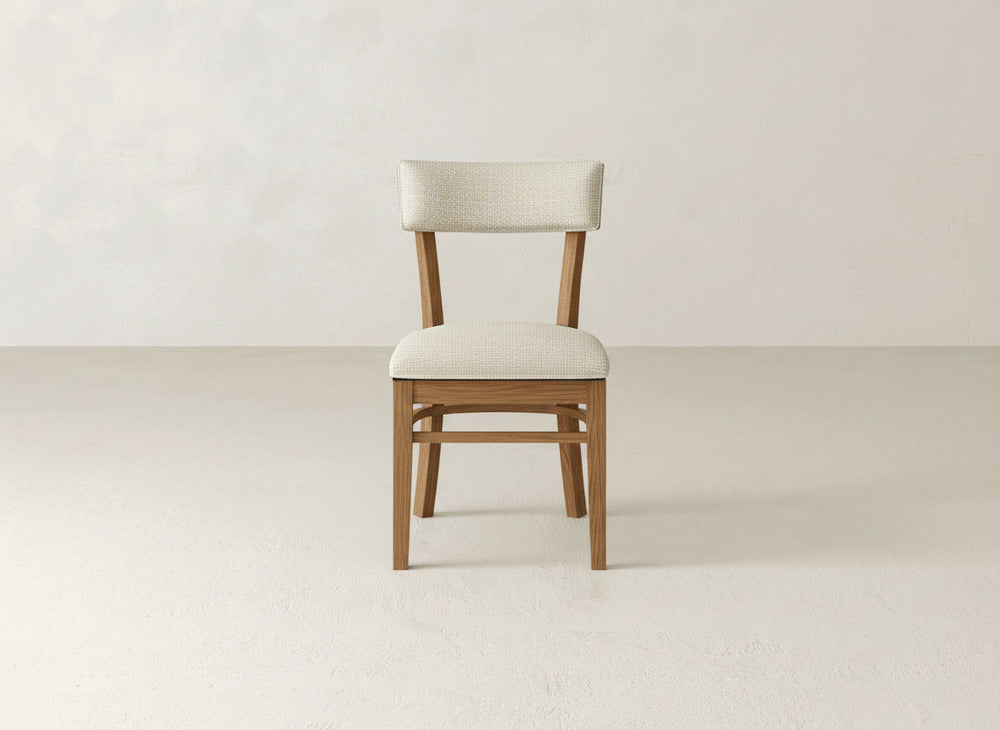 The Everly Side Dining Chair by James+James, featuring a light beige upholstered seat and backrest, is showcased in a minimalist room with off-white walls and a light floor.