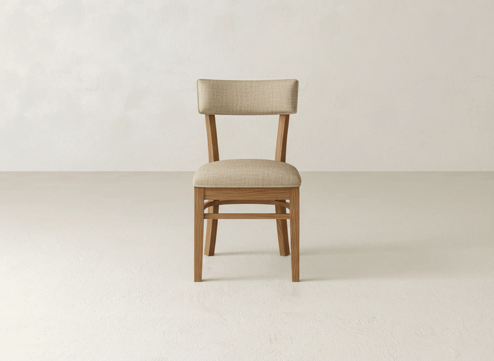 The Everly Side Dining Chair by James+James features a simple, modern wooden frame with padded beige seat and backrest, shown on a light floor against a neutral textured wall.