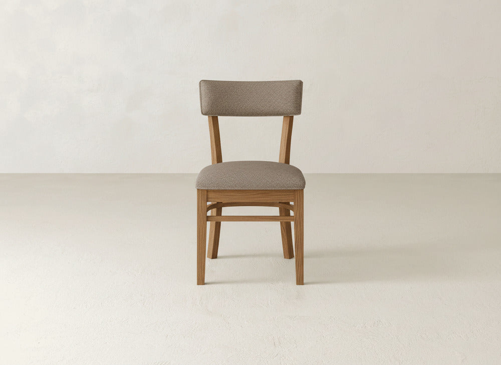The Everly Side Dining Chair by James+James, featuring a light brown cushioned seat and backrest with wood construction, is shown facing forward against a plain off-white wall and floor.