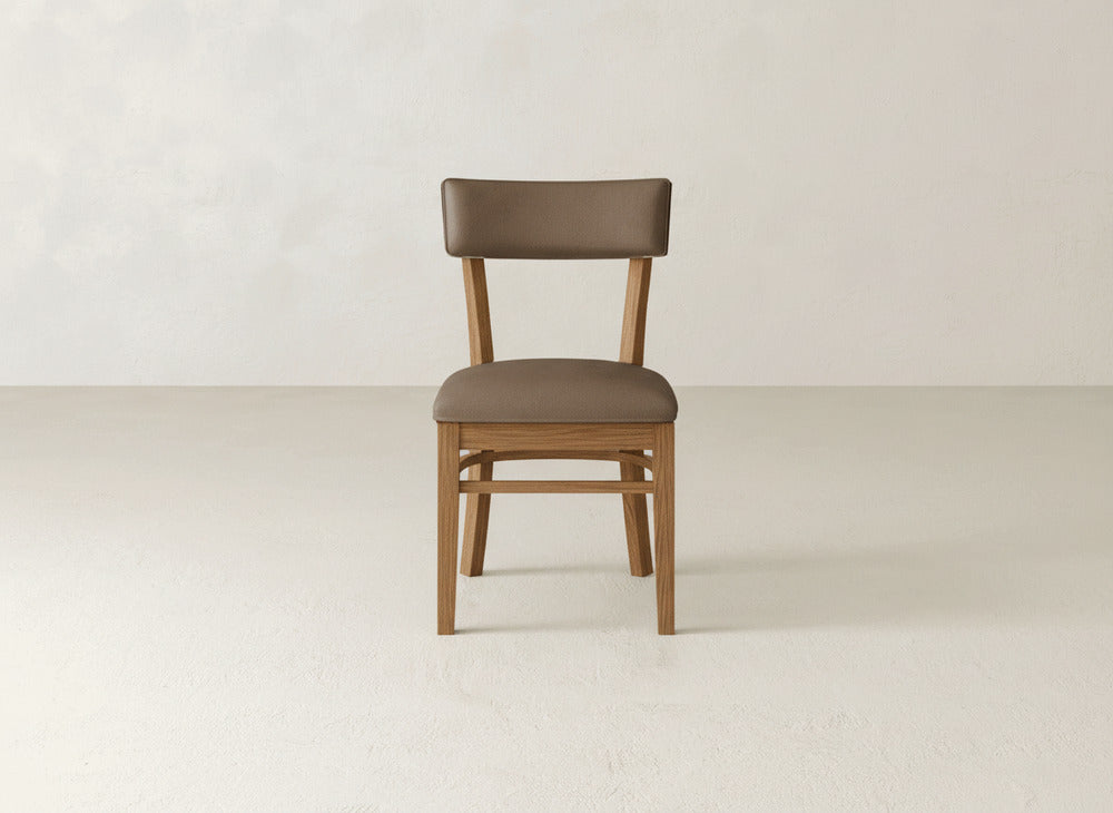 The Everly Side Dining Chair by James+James, featuring a brown upholstered seat and backrest, is centered in an empty, light-colored room with plain walls and flooring.
