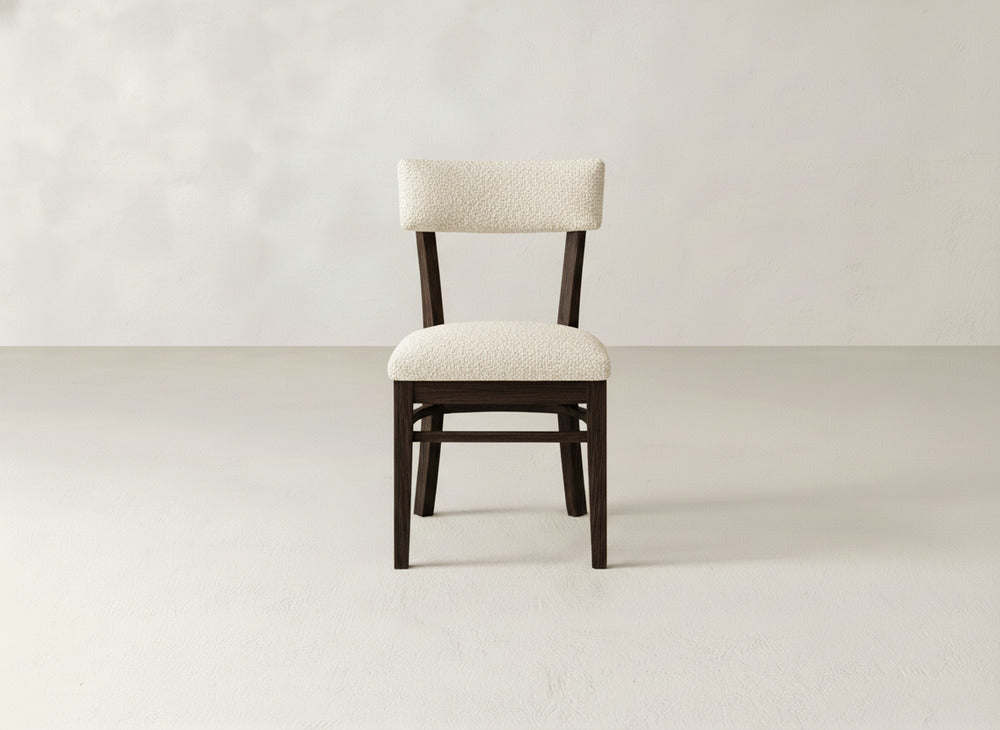 The Everly Side Dining Chair by James+James features dark wooden legs and a light, textured upholstered seat and backrest, perfectly centered in a minimalist room with a plain, light-colored wall and floor.