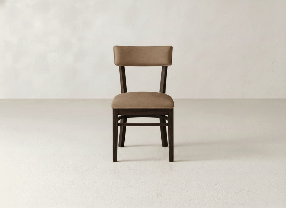 The Everly Side Dining Chair by James+James, featuring an upholstered seat and backrest, stands centered on a light floor against an off-white wall. The brown wood chair is positioned facing forward in the minimalist space.