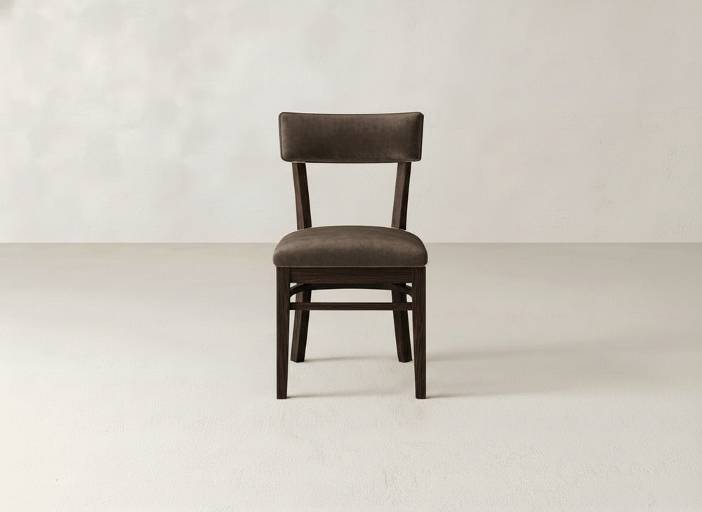 The Everly Side Dining Chair by James+James, featuring a dark wood frame and brown upholstered seat and backrest, sits centered on a light-colored floor against an off-white wall. Its simple, modern design showcases clean lines.