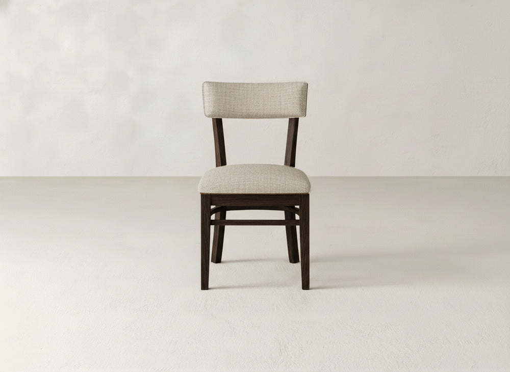 The Everly Side Dining Chair by James+James, featuring a light beige upholstered seat and backrest, is centered in a minimal, empty room with light floors and walls.