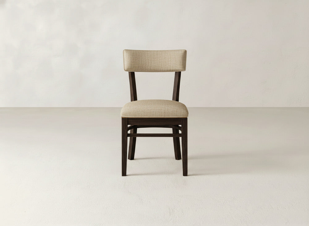 The Everly Side Dining Chair by James+James features a light beige upholstered seat and backrest with a simple wooden frame, placed centrally in a plain room with light-colored flooring and walls.
