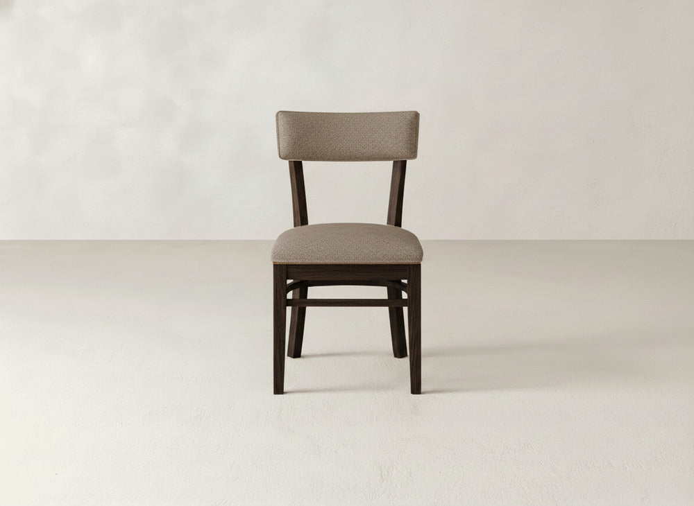 The James+James Everly Side Dining Chair, featuring a light brown upholstered seat and backrest, stands alone in the center of an empty room with light-colored walls and floor.
