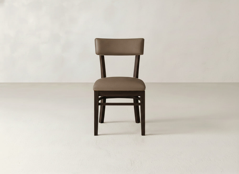 The Everly Side Dining Chair by James+James, featuring a padded brown upholstered seat and backrest, is centered on a light-colored floor against an off-white wall and photographed from the front.