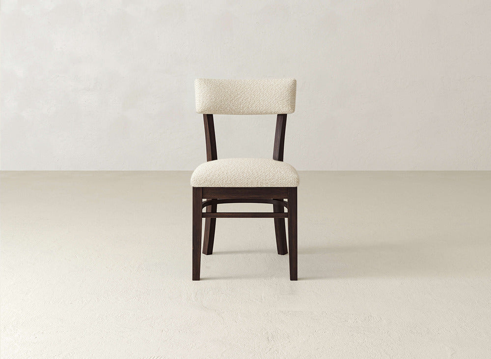 The Everly Side Dining Chair by James+James features dark wooden legs and a light, textured upholstered seat and backrest, shown on a plain, light-colored floor against a neutral wall.