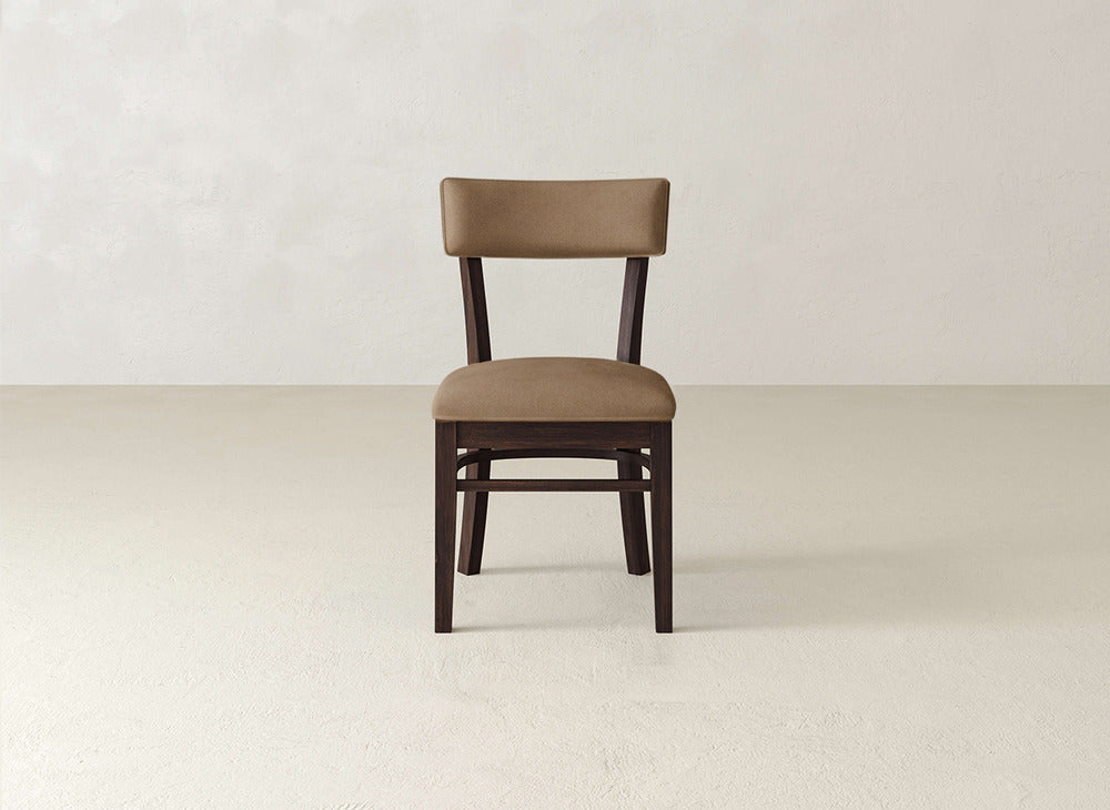 The Everly Side Dining Chair by James+James features a wooden frame and cushioned seat and backrest upholstered in brown fabric, set on a light floor against an off-white wall.