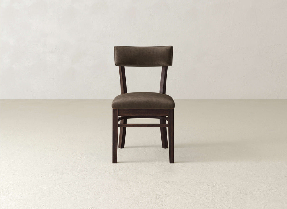 The Everly Side Dining Chair by James+James features a simple mid-century design with a dark brown cushioned seat and backrest, set on a light floor against an off-white wall.