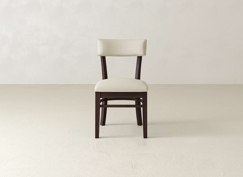 The Everly Side Dining Chair by James+James features a cushioned seat and backrest in light beige fabric, elegantly set against a plain, light-colored wall and floor background.