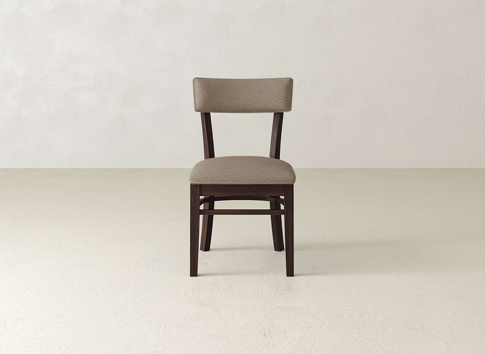 The Everly Side Dining Chair by James+James features a wooden frame with cushioned seat and backrest in light brown fabric, set on a plain, light-colored floor against a neutral wall.