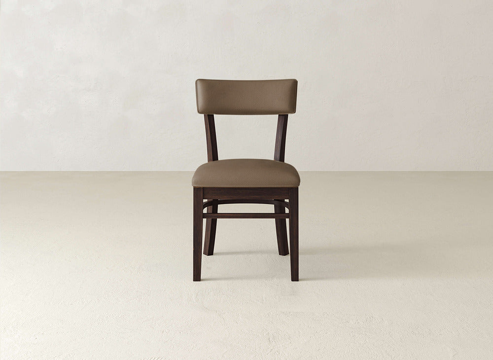 The Everly Side Dining Chair by James+James features a wooden frame with a brown padded seat and backrest, displayed against a plain light-colored wall and floor.