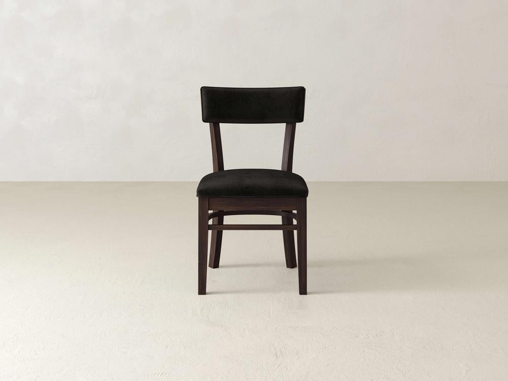 A simple, dark wooden chair with a black cushioned seat and backrest stands on a light, plain floor against an off-white wall. The chair has a minimalist design with straight legs and a rectangular backrest.