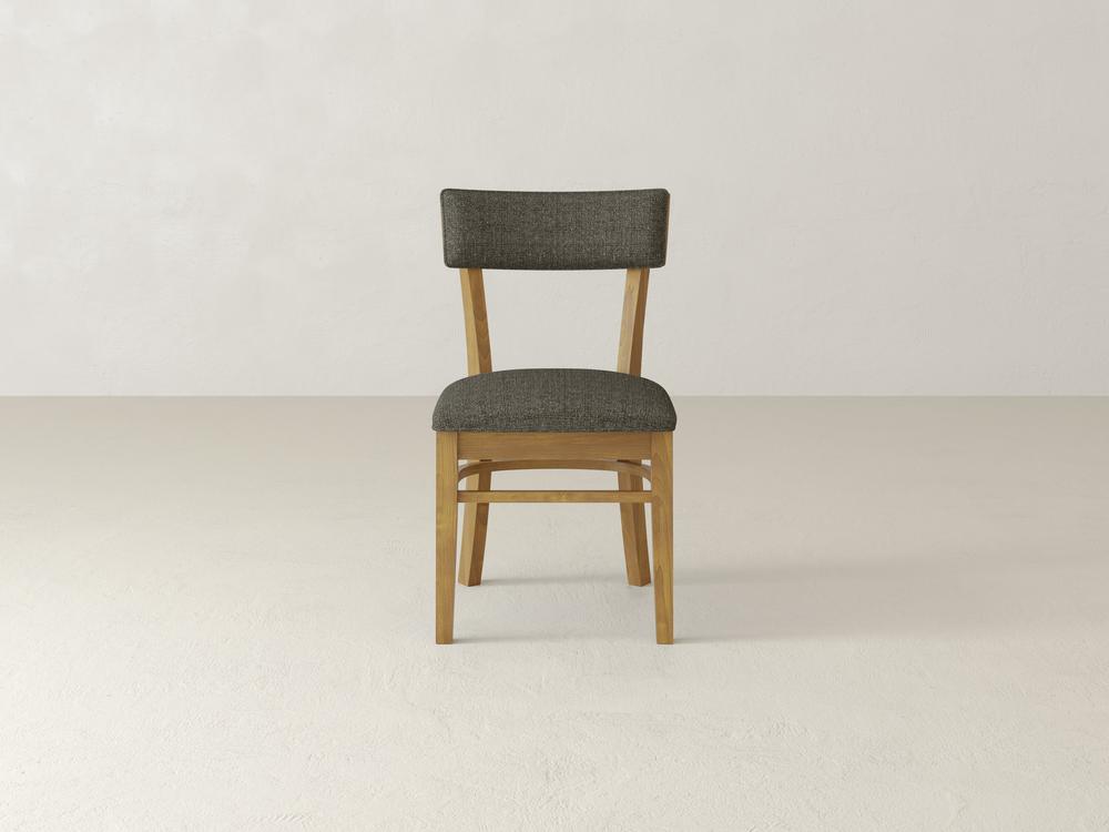A wooden chair with a dark gray cushioned seat and backrest is placed in the center of a light, minimalist room with a pale floor and wall.