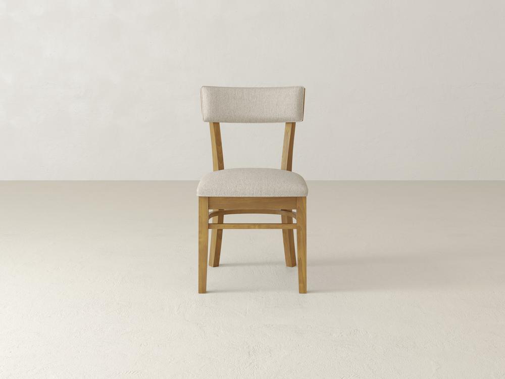 A wooden chair with a light beige cushioned seat and backrest, positioned against a plain off-white background. The chair has simple, straight lines and a minimalist design.