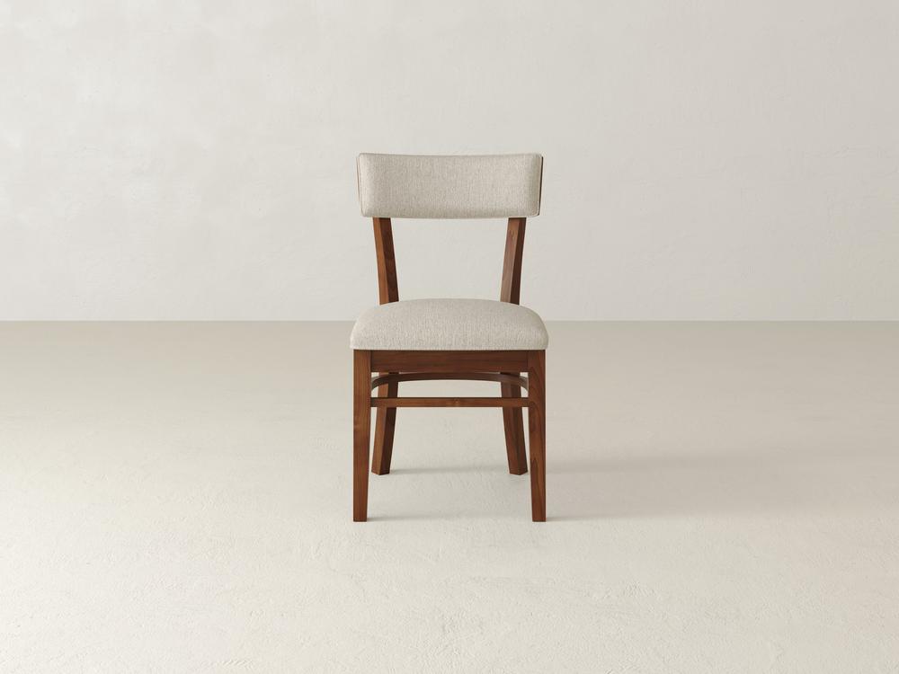 A wooden chair with a cushioned beige fabric seat and backrest is positioned on a plain, light-colored floor against a neutral backdrop. The chair has a simple, classic design.