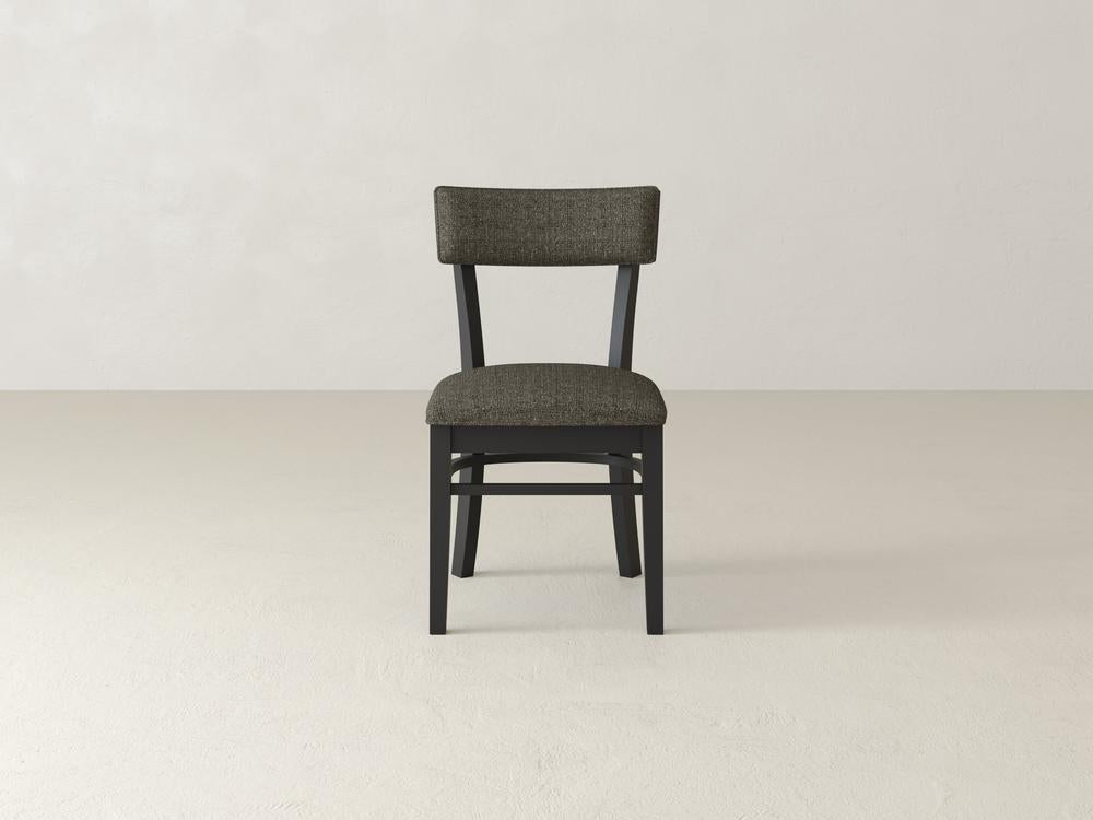 A dark grey upholstered chair with a wooden frame stands on a light-colored, textured floor against a plain, off-white wall.