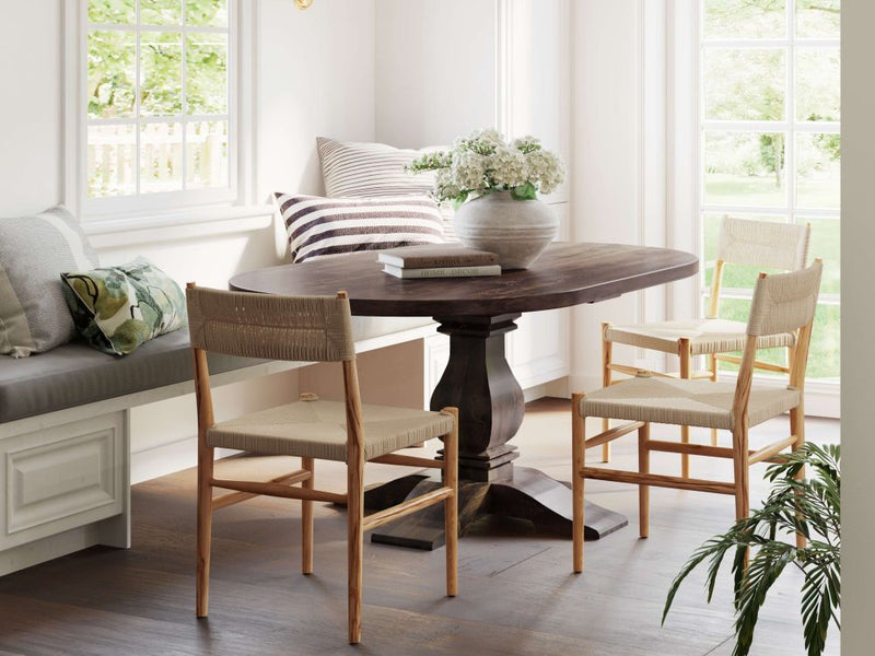 A cozy corner dining area highlights the James & James Expandable Round Heirloom Dining Table with a knotty alder tabletop, surrounded by four woven chairs and a cushioned bench with striped pillows. A white flower vase adorns the table as a green plant below adds life, all bathed in bright natural light.
