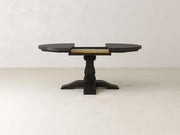 A dark wooden pedestal table is shown with its round top partially extended. The table has a central column and a sturdy base. It stands on a plain light-colored floor against a neutral wall.