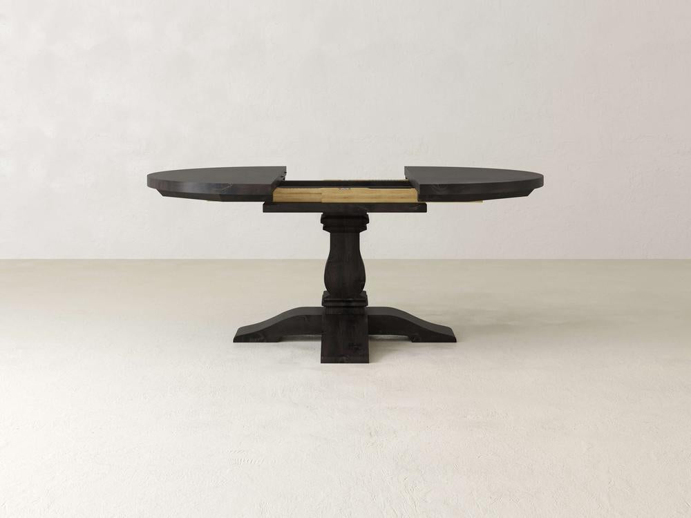 A dark wooden pedestal table is shown with its round top partially extended. The table has a central column and a sturdy base. It stands on a plain light-colored floor against a neutral wall.