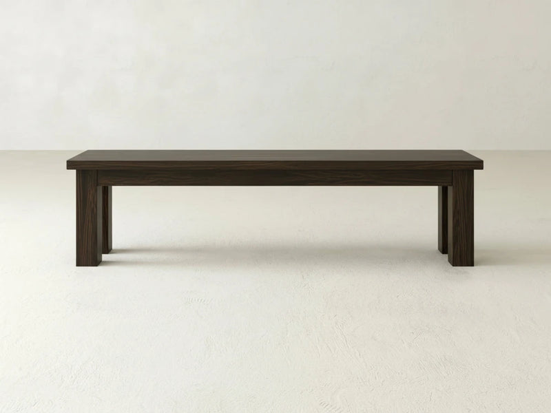 The James+James Farmhouse Bench in Espresso on Oak features a simple, rectangular design and sits on a light-colored floor against an off-white wall.