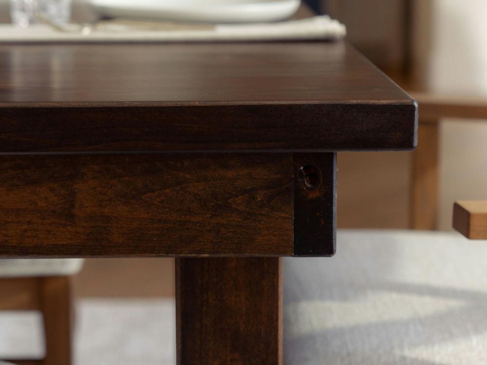 Close-up of the corner of a Farmhouse Dining Table - Tobacco by James+James. Part of a wooden chair with a light-colored cushion can be seen beneath the table. The background shows a blurred section of another chair and a dining mat with plates and cutlery on the tabletop.