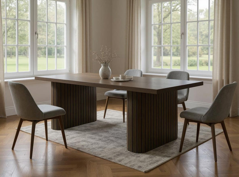 A modern dining room features the James+James Fern Dining Table in Charred Ember, four upholstered chairs, a beige rug, and a vase with dried branches. Large windows with cream curtains fill the space with natural light and overlook a green lawn.