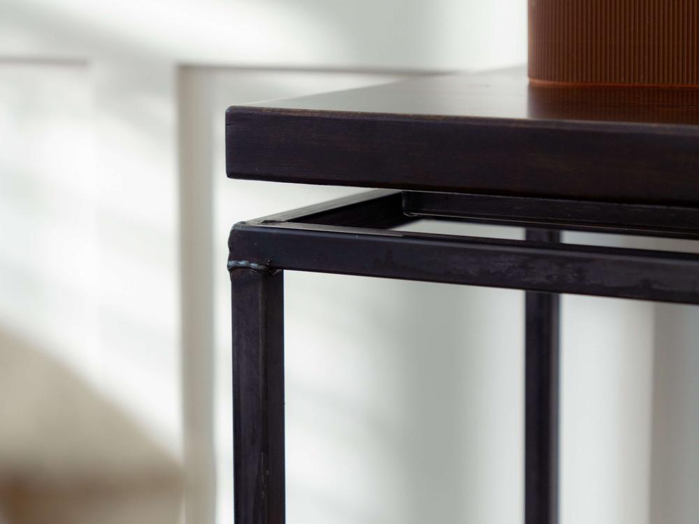 A close-up of a James+James Floating Top Sofa Table - Tobacco corner. The metal legs have a sleek, black finish, and the dark wooden tabletop is polished. Bright, natural light casts shadows on the table from a nearby window, emphasizing the clean, angular lines of the design.