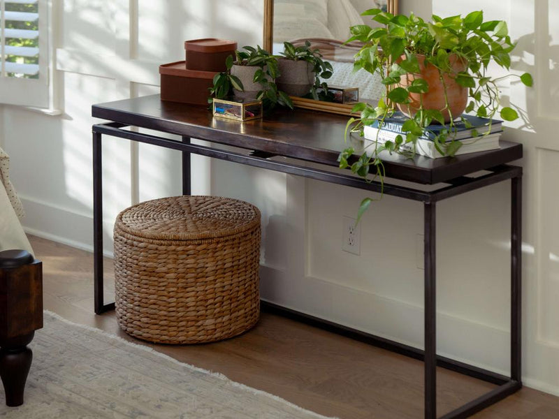 The Floating Top Sofa Table - Tobacco by James+James, with its sleek black metal frame and rich dark wooden top, stands against a white wall. On it sit potted plants, a stack of books, and decorative boxes. Beneath the table is a round woven ottoman. A large mirror on the table reflects part of the room.