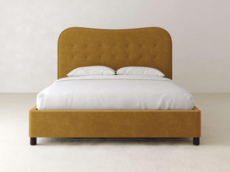 The Florence Bed – Velvet by James & James features a mustard-colored velvet upholstery and high tufted headboard, set against a light wall. It combines modern timeless appeal with white bedding and two pillows, resting elegantly on solid maple feet on a light-colored floor.