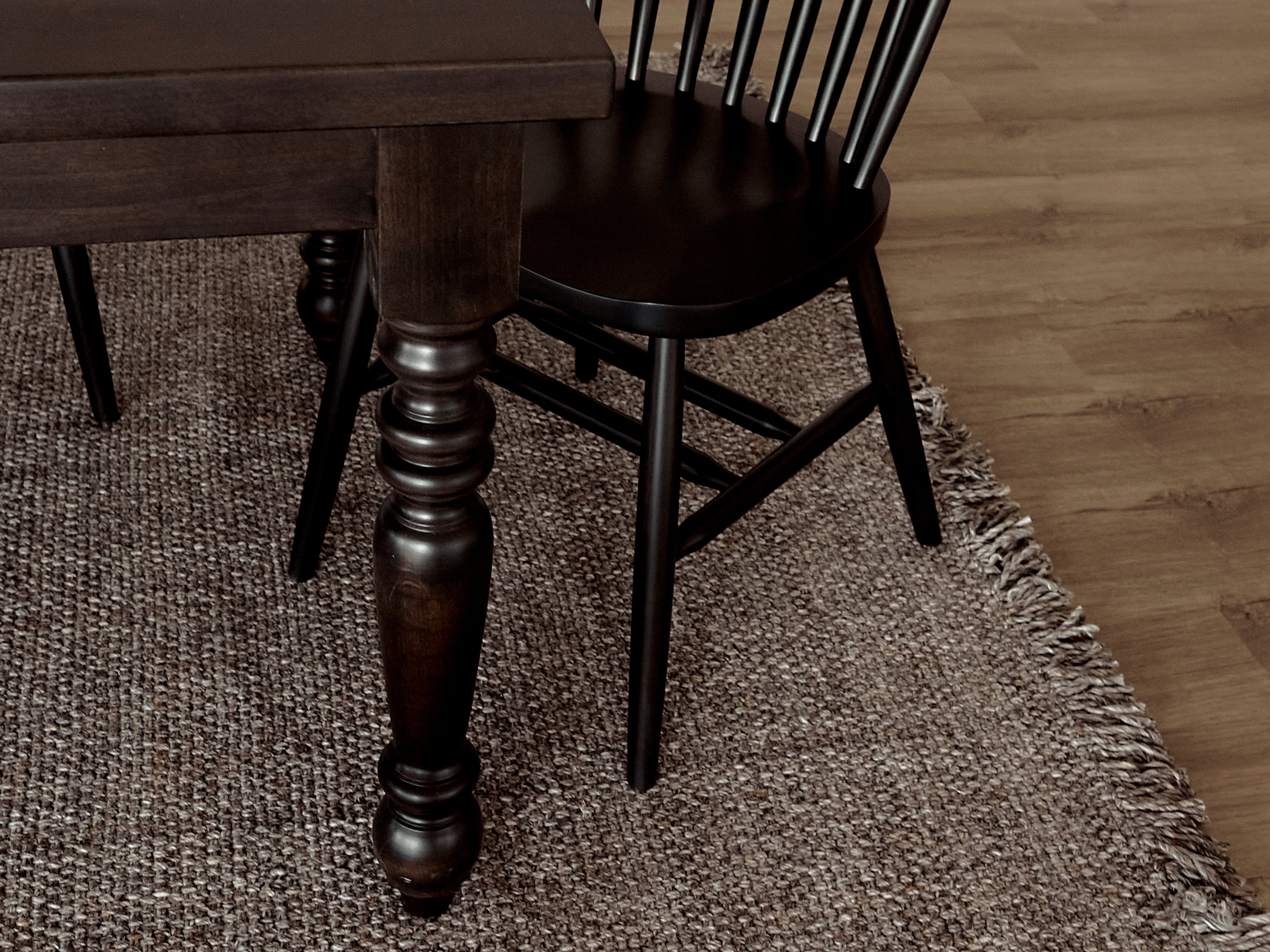 A James+James French Country Dining Table in Harvest Wheat stands beside a black spindle chair, both set atop a textured woven rug on a wooden floor.