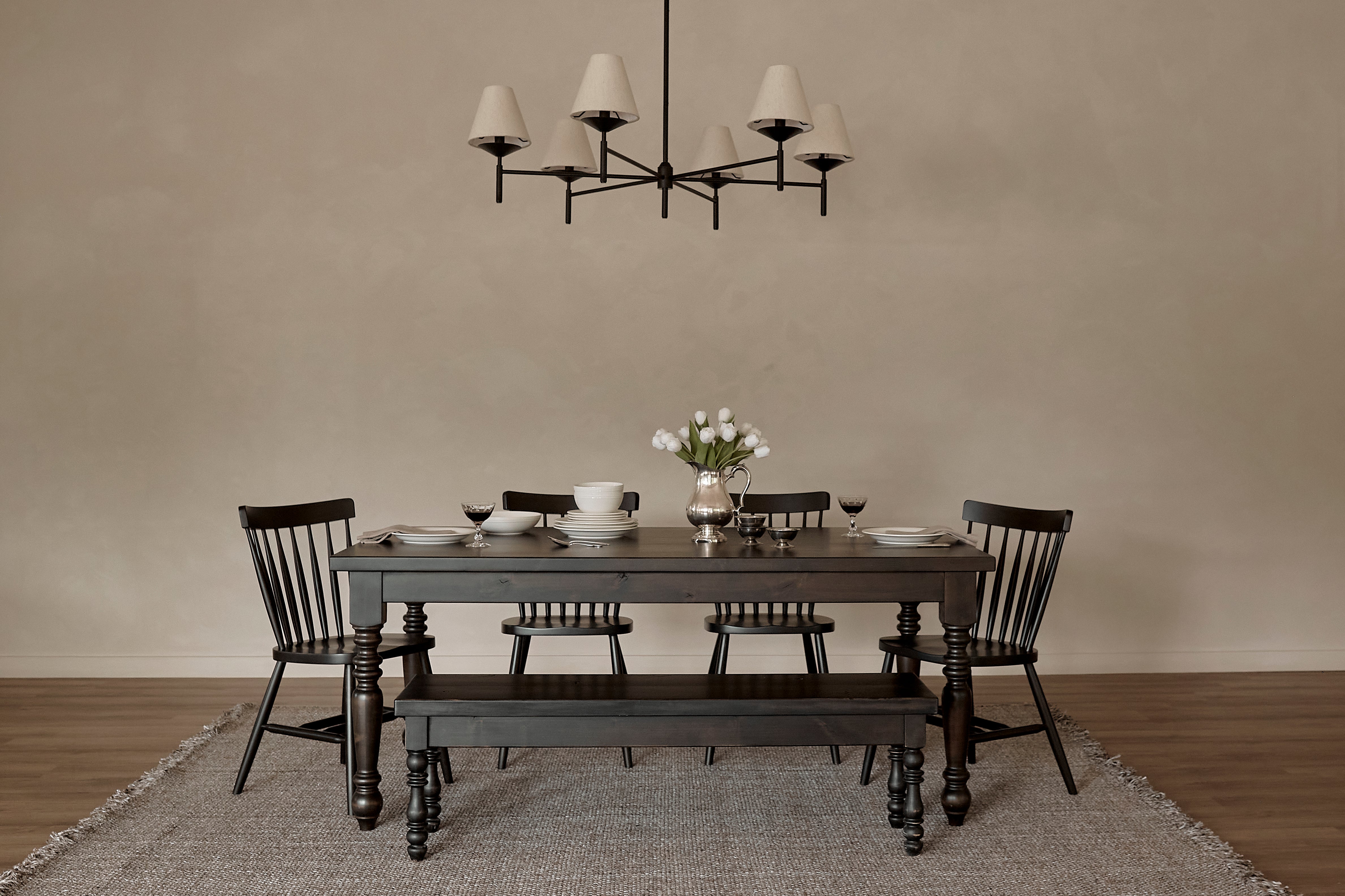 A minimalist dining room features the James+James French Country Dining Table in Harvest Wheat, four matching chairs, a bench, set with dishes and a vase of white flowers. A modern chandelier hangs above, with a neutral rug on the wooden floor.