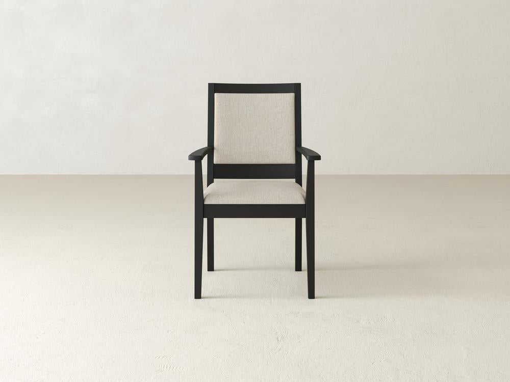 A wooden chair with a beige cushion sits on a light-colored floor against a plain white wall. The chair has a dark frame and armrests, with a minimalist design.