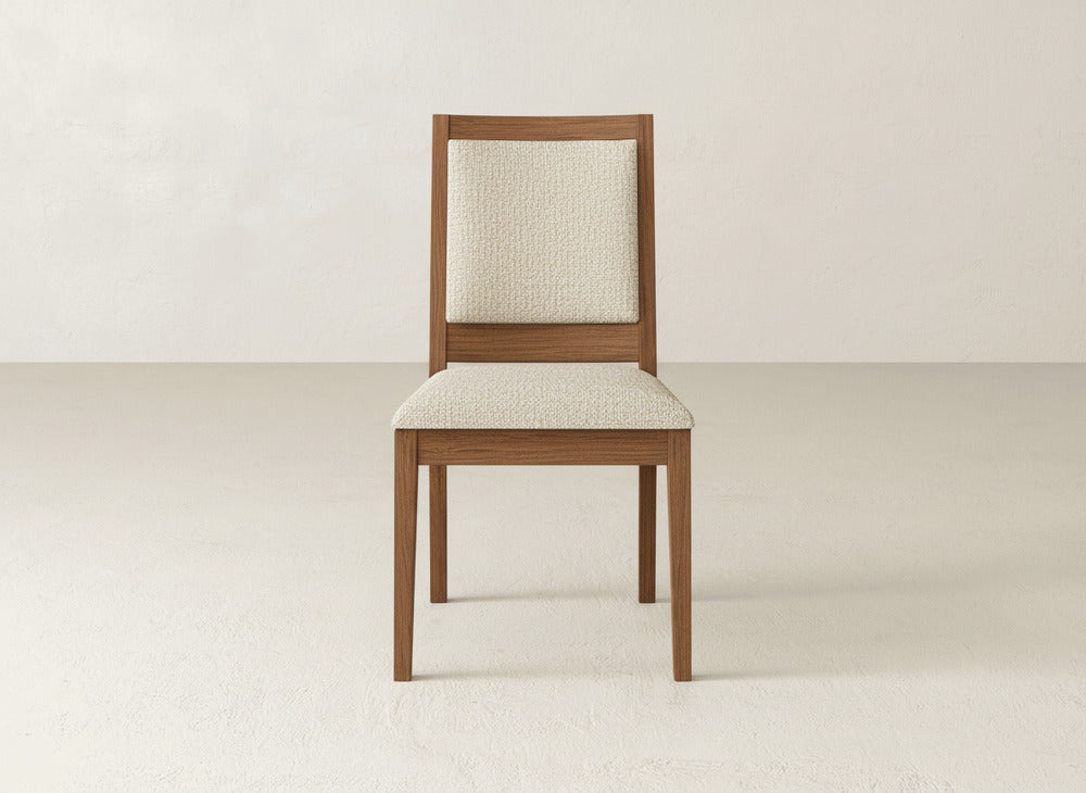 The Gina Side Dining Chair by James+James, featuring maple wood and a light beige upholstered seat and backrest, is set against a plain, light-colored wall and floor.