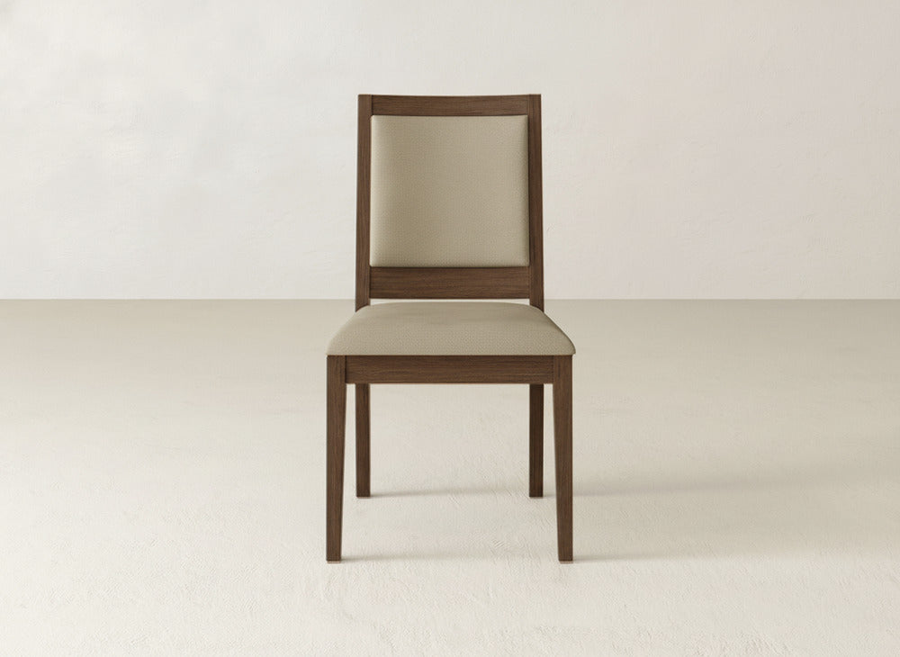 A James+James Gina Side Dining Chair with a maple wood frame and beige cushioned seat and backrest is centered against a plain, light-colored wall and floor.