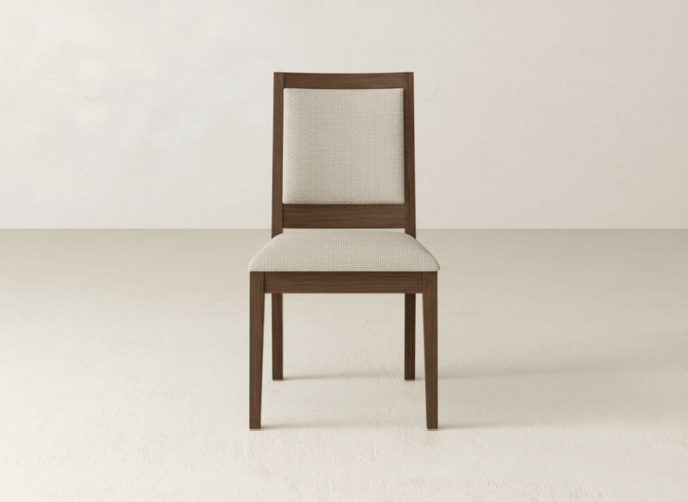 The James+James Gina Side Dining Chair, featuring a maple wood frame and light beige upholstered seat and backrest, is positioned against a plain, light-toned wall and floor.