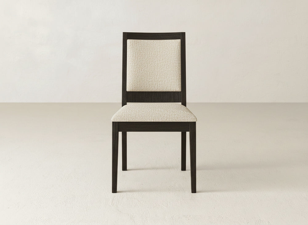 A James+James Gina Side Dining Chair, featuring a dark maple frame and light upholstered seat and backrest, stands in the center of a minimalist, light-toned room.