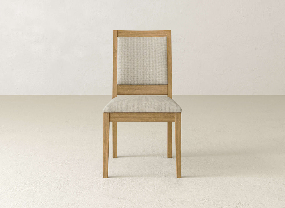 The James+James Gina Side Dining Chair features maple wood and a light beige upholstered seat and backrest, shown facing forward against a plain off-white wall and floor background.
